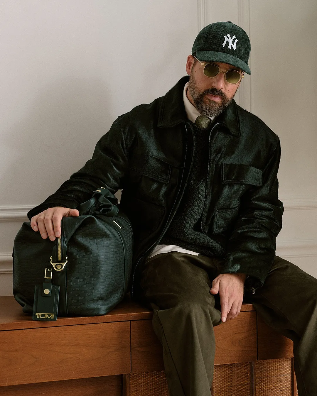 Man in a green leather jacket and cap sitting beside a leather duffle bag